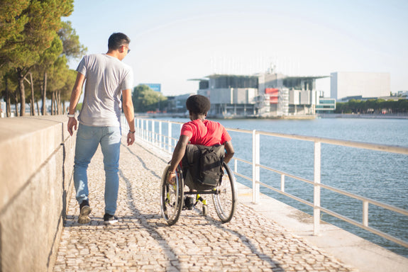 Dos amigos paseando de espaldas por un paseo empedrado junto al agua, uno caminando y el otro en silla de ruedas manual, con edificios contemporáneos al fondo.