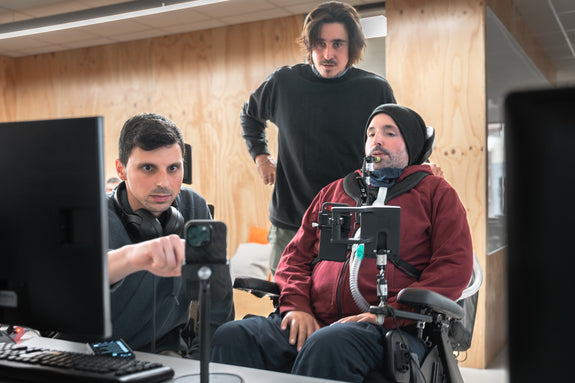 Three men collaborating at a computer workstation. One of them, using a power wheelchair and a device (MouthX), controls the screen through assistive technology.